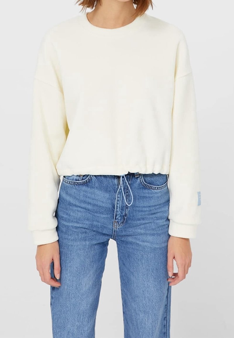 Femme portant un sweat-shirt court à manches longues couleur écru avec un cordon de serrage à l'ourlet et un jean bleu taille haute, debout devant un fond uni.