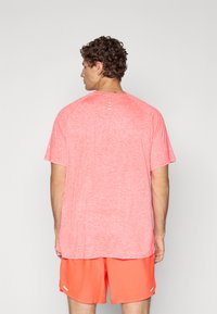Hombre con cabello rizado que lleva una camiseta deportiva de manga corta en un brillante color coral y pantalones cortos a juego, de espaldas frente a un fondo plano.