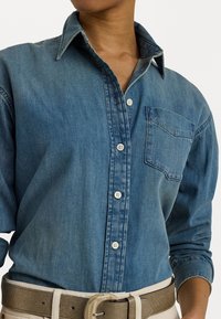 Košile z denimu s knoflíky v světlé modré barvě, obsahující límec, náprsní kapsu a bílé knoflíky. Tkanina má měkký, strukturovaný vzhled.