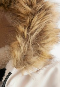 Bordure en fausse fourrure dans des nuances de brun clair et de beige, texture douce se superposant à un tissu blanc avec un détail de fermeture éclair noir.