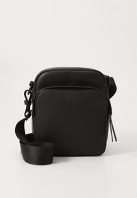 Pieni musta olkalaukku, jossa on etutasku vetoketjulla ja säädettävä olkahihna, valmistettu sileästä nahkaa muistuttavasta materiaalista yksivärisellä taustalla.