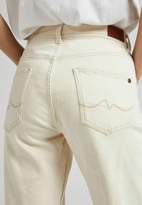 Lähikuva henkilöstä, jolla on päällä vaaleankermanväriset korkeavyötäröiset farkut, joiden takataskuissa on ommeltu aalto-tyyppinen kuvio, sekä valkoinen paidanhelma sisään laitettuna.