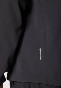 Schwarze Windbreaker-Jacke mit glatter Textur, sleekem Design und sichtbarem "WINDWALL"-Logo an der Seite. Maßgeschneiderte Passform mit minimalen Nähten.