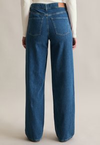 Osoba nosząca niebieskie dżinsy z wysokim stanem i szerokimi nogawkami, z tylnymi kieszeniami oraz beżową skórzaną metką, stojąca na jednolitym tle.