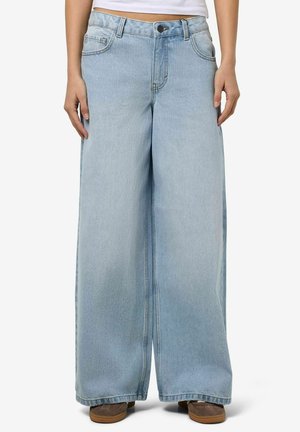 Jasnoniebieskie dżinsy z wysokim stanem i szerokimi nogawkami noszone przez osobę z rękami swobodnie opuszczonymi wzdłuż ciała, a pod krawędzią widoczne brązowe buty.