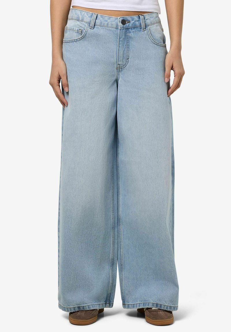 Jean large taille haute bleu clair porté par une personne avec les mains détendues le long du corps et des chaussures marron visibles sous l'ourlet.