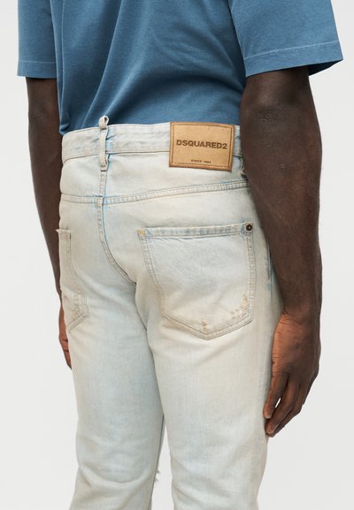 Homme portant un jean délavé bleu clair avec un patch en cuir de la marque DSQUARED2 à l'arrière, assorti à une chemise à manches courtes bleu moyen.