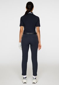 Donna che indossa una polo blu navy, pantaloni blu fitted, guanto da golf bianco e sneakers bianche, in piedi di spalle contro uno sfondo neutro.
