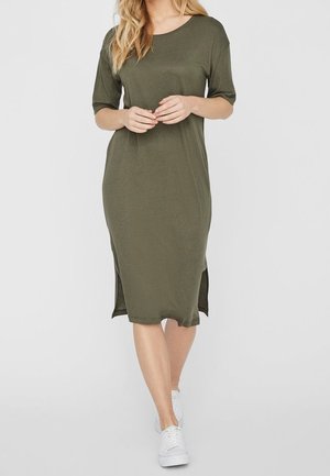 Kobieta w oliwkowej, midi sukience z rękawami do łokcia i rozcięciami po bokach, w białych trampkach, idąca do przodu.