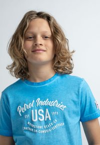 Blå bomullströja med rund halsringning, försedd med vittryckt text: "Petrol Industries USA 1973" och en logodetalj på ärmen.