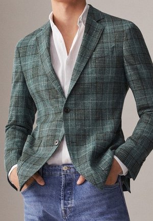 Mand iført en grøn ternet blazer over en åben hvid skjorte med hænderne i lommerne på blå jeans.