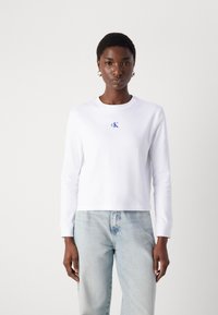 Maglietta bianca a maniche lunghe con collo rotondo, caratterizzata da un logo ricamato "CK" blu sul petto. Tessuto morbido e liscio, design leggermente accorciato.