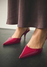 Escarpins slingback à bout pointu, brillant, de couleur rose vif avec un talon stiletto fin et un design découpé sur les côtés, posés sur une surface sombre.