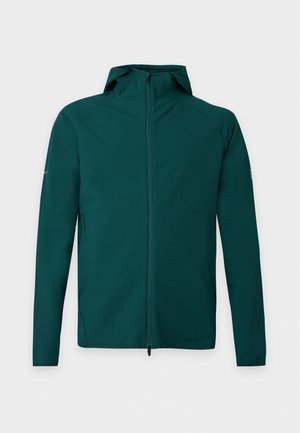Homme portant une veste à capuche verte, un t-shirt blanc, un jean bleu foncé et des baskets blanches, debout devant un fond uni.