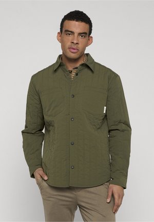 Uomo che indossa una giacca trapuntata verde oliva con bottoni a pressione e pantaloni beige, in piedi con una mano in tasca contro uno sfondo chiaro.