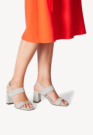 Light gray block-heel sandals with adjustable ankle straps, featuring a gold accent detail. Worn with an orange dress, showing toes.
