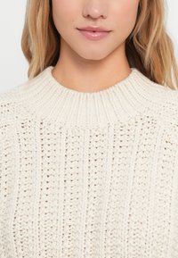 Knitted cream sweater with a high ribbed collar and vertical stripe texture. Fabric appears soft and slightly chunky.