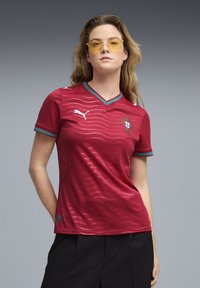 Κόκκινη ποδοσφαιρική φανέλα με κοντά μανίκια και V-λαιμόκοψη, με υφασμάτινο ριγέ μοτίβο, πράσινες λεπτομέρειες και το εθνόσημο της Πορτογαλίας.