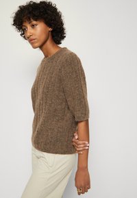 Donna con i capelli ricci che indossa un maglione corto a maniche corte di colore marrone texture e pantaloni crema, mentre tiene il braccio sinistro con la mano destra.