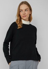 Schwarzer Strickpullover mit Rundhalsausschnitt, gerippten Bündchen und Saum. Wird über einem weißen Rollkragenpullover mit lockerem Schnitt und weicher Textur getragen.