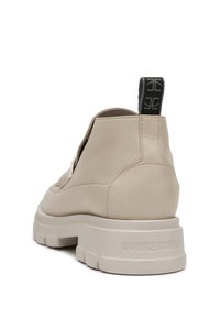 Zapatos de cuero beige con suela gruesa y acabado texturizado. Presentan un lazo negro en el talón y un sutil logo en el costado.