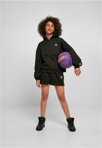 Conjunto de sudadera oversized negra y pantalones cortos de algodón, con un pequeño parche de logo. El modelo sostiene una pelota de baloncesto morada y lleva zapatillas altas negras.