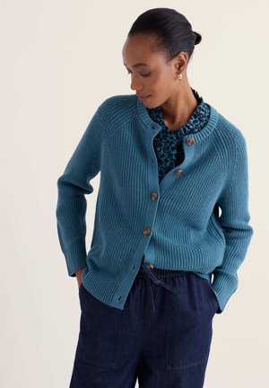 Femme portant un cardigan en tricot côtelé bleu sarcelle avec des boutons marron par-dessus un chemisier bleu à motifs et un pantalon bleu foncé, regardant vers le bas, les mains dans les poches.