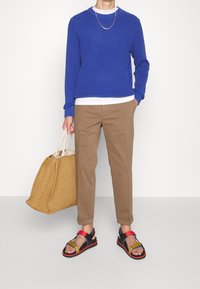 Pull bleu côtelé, t-shirt blanc, pantalon beige, sandales multicolores, tenant un sac fourre-tout en paille tissée. Tenue simple et décontractée.