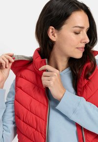 Rød polstret vest med høj krave og lynlås, der har et let design og quiltet tekstur, båret over en lyseblå sweater.