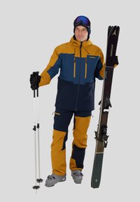 Équipement de ski comprenant une veste isolante orange et bleue, des gants noirs, un pantalon marine, des chaussures de ski grises et des skis avec des fixations noires.