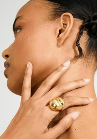 Woman with smooth skin and braided hair touches her neck, wearing a large, shiny, gold, heart-shaped ring on her finger.