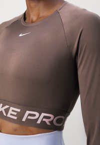 Nahaufnahme einer Person, die ein braunes Nike Pro Langarm-Crop-Top mit einem weißen Swoosh-Logo auf der Brust trägt.
