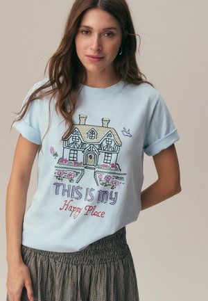 Frau mit langen braunen Haaren trägt hellblaues, hochgekrempeltes Hemd mit einer Stickerei eines Hauses, Blumen und dem Text „Dies ist mein glücklicher Ort.“