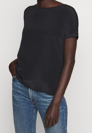 Czarny top z krótkim rękawem wykonany z gładkiego materiału, z okrągłym dekoltem i luźnym krojem, w zestawieniu z jasnoniebieskimi dżinsami.