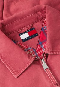 Col de veste rouge avec fermeture éclair argentée, doté d'une doublure intérieure à carreaux rouge et bleu et d'une étiquette logo Tommy Jeans à l'intérieur de l'encolure.