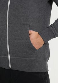 Grå glidelåshette laget av bomullsmiks, med sidelommer, ribbede mansjetter og en hvit glidelås. Jevn tekstur med et uformelt design.