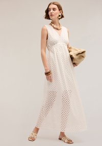 Rochie maxi albă fără mâneci, cu model perforat, decolteu în V, talie strânsă și suprapose din material transparent. Este însoțită de o poșetă bej și sandale.