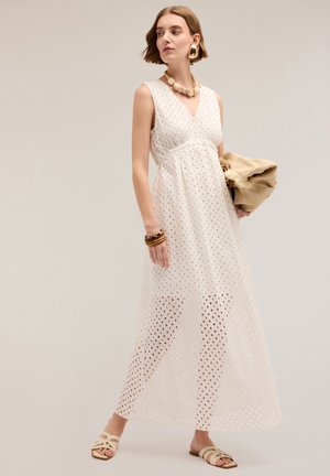 Witte mouwloze maxi-jurk met gaatjespatroon, V-hals, cinched taille en doorzichtige overlay. Vergezeld van een beige tas en sandalen.