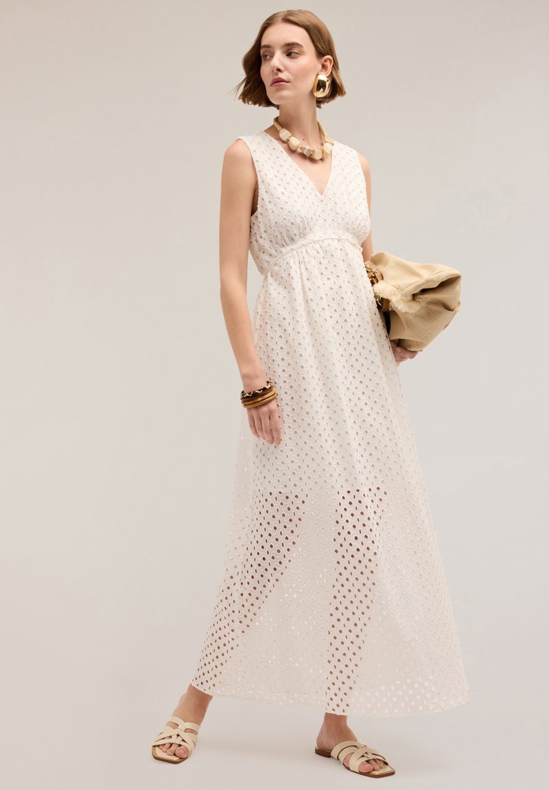 Rochie maxi albă fără mâneci, cu model perforat, decolteu în V, talie strânsă și suprapose din material transparent. Este însoțită de o poșetă bej și sandale.