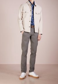 Béžová džínová bunda přes tmavě modrou košili, doplněná šedými kalhotami a bílými teniskami s hnědými podrážkami. Bunda má dvě kapsy.