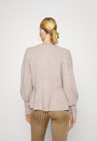 Mujer con coleta rubia, que lleva pendientes de aro, un suéter de punto ligero en color malva con mangas abullonadas y pantalones marrones con rayas verticales, de espaldas.