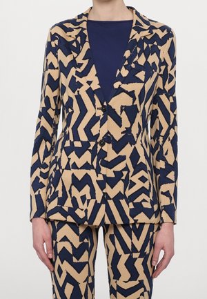 Personne portant un blazer aux motifs abstraits en zigzag beige et bleu marine, avec un pantalon assorti, et un haut uni bleu marine en dessous.