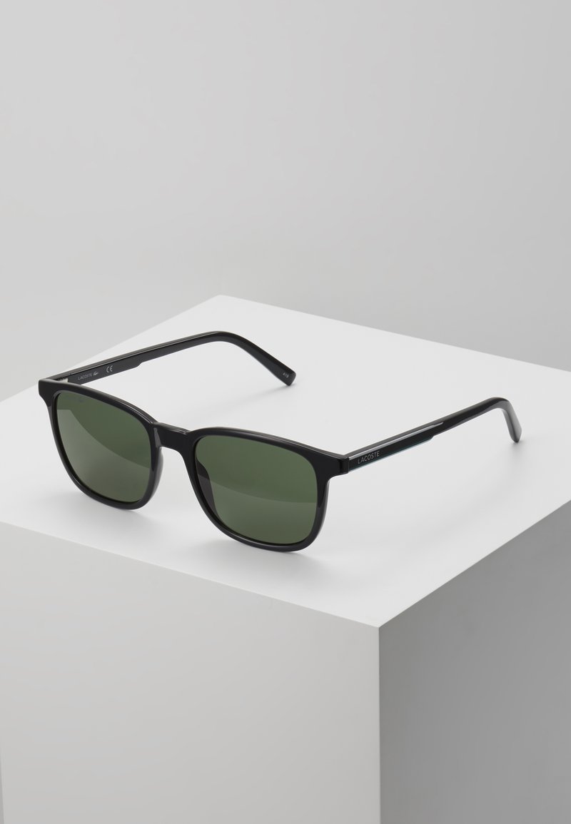 Ochelari de soare negri cu lentile de un verde închis, rame rectangulare și brațe subțiri, cu un detaliu subtil de marcă pe lateral.