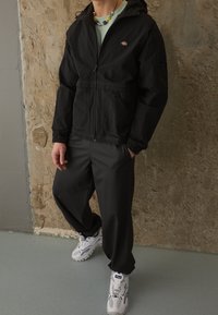 Svart zip-up jacka med justerbar huva, elastiska manschetter och ficka. Grå, löst sittande byxor. Vita sneakers med svarta detaljer.