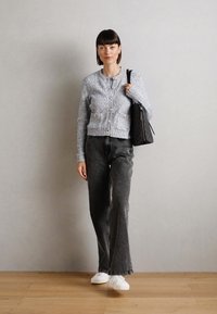 Cardigan di maglia grigio chiaro con bottoni, tasche frontali, abbinato a jeans grigi scuri a zampa d'elefante e sneakers bianche, con borsa nera tenuta sul lato sinistro.