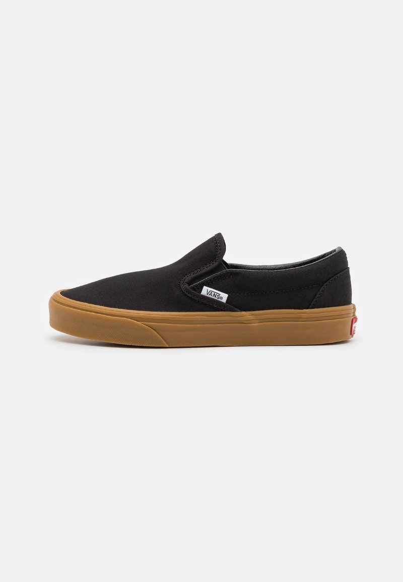 Mustat slip-on Vans -tossut, joissa on ruskea kumipohja, pieni valkoinen Vans-logo-tagi sivussa ja punainen kantapäälogo valkoisella taustalla.