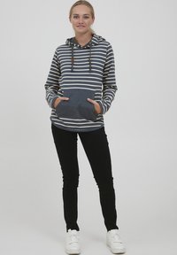 Sudadera a rayas en navy e marfil, con un bolsillo frontal y capucha con cordón. Combinada con pantalones negros ajustados y zapatillas blancas.