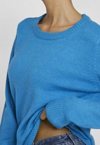 Pull en maille bleu avec un col rond, des poignets et un ourlet côtelés. Le tissu semble doux, avec une texture lisse et une coupe légèrement ample.