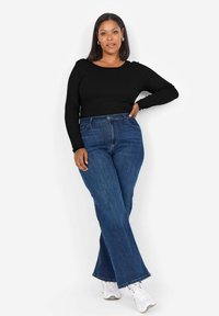 Svart långärmad topp som matchas med högmidjade blå denimflare jeans. Modellen bär vita sneakers och visar en avslappnad, figurnära stil.