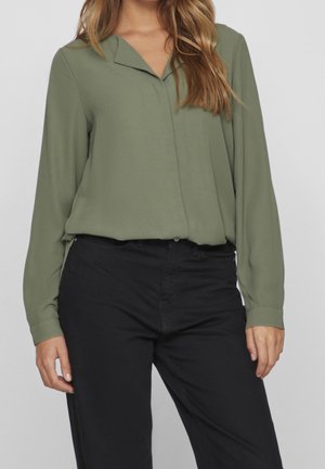 Blusa verde oliva a maniche lunghe con colletto a V, in tessuto morbido e chiusura con bottoni. Abbinata a pantaloni neri a vita alta.